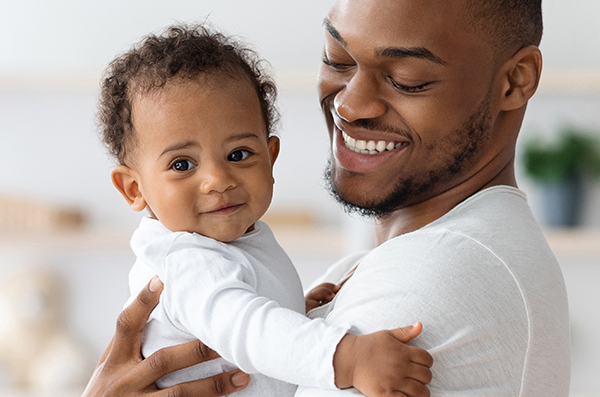 A smiling man holds a happy baby in his arms. Both are wearing white shirts. The background is softly blurred, creating a warm and intimate atmosphere.