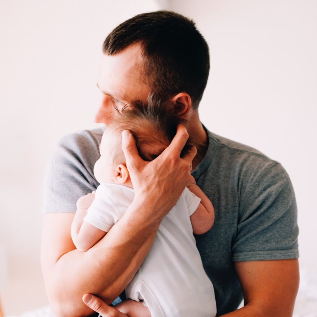 A man in a gray t-shirt gently holds a baby wearing a white onesie. The man is looking away from the camera, and the baby is resting on his chest with eyes closed. The background is softly blurred.