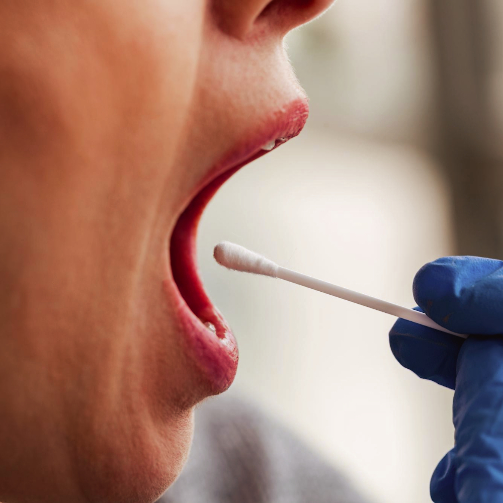 A close-up of a person having their mouth swabbed. The person has their mouth open, and a gloved hand holds a cotton swab near their cheek, likely for a medical test.