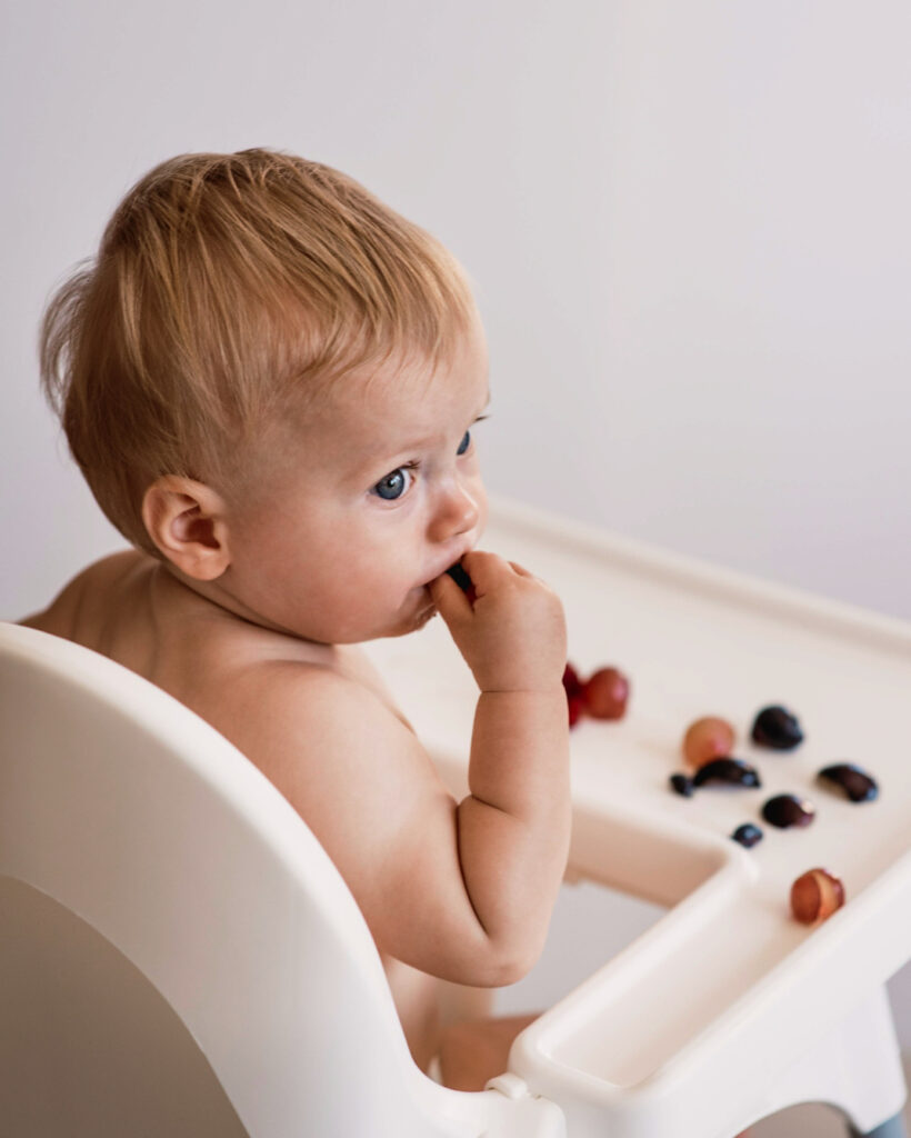 A baby with blonde hair sits in a high chair, eating berries from a tray. The background is plain, and the baby is focused on the fruit.